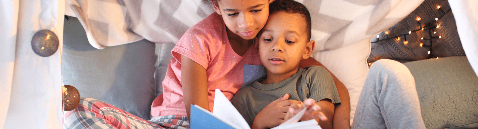 Two children reading a book in a fort made of pillows and blankets